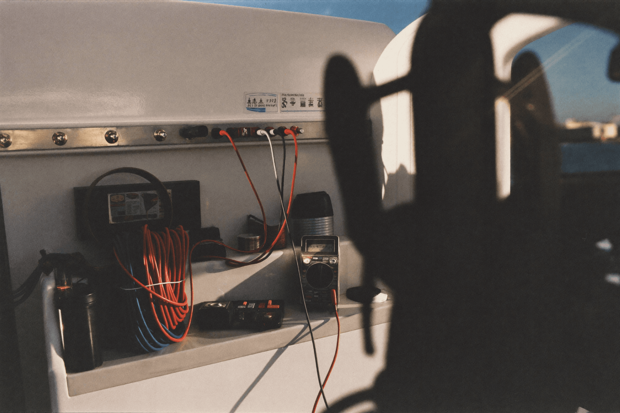 Multimeter probes on a boat's battery bus bar beside an open marine electrical panel and coils of tinned cable.