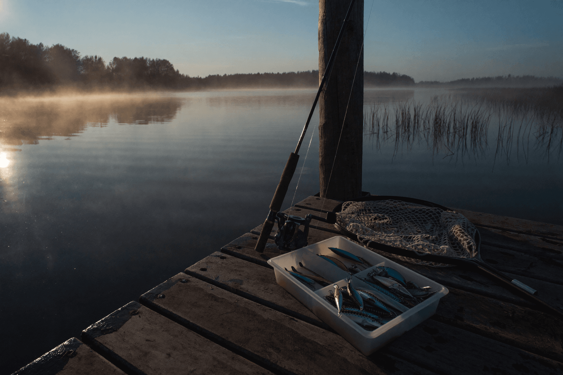 Spinning rod, open lure tray and rubber landing net on a wooden dock at sunrise over a misty lake